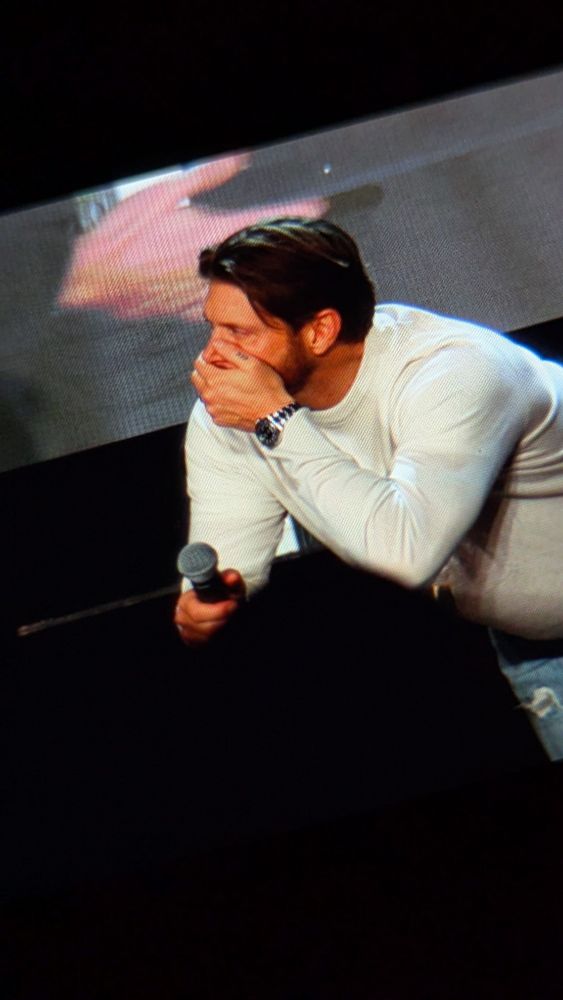 Jensen leaming on the back of the chair while standing behind it, hand on his mouth while looking at Misha off frame to the left.