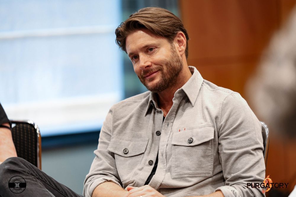 Jensen is sitting on a chair visible midsection up. He is facing to the left of the camera and is looking and listening to someone in the same direction. He has a slight smile on his face. You can see his right knee, which is a bit on the side while leaning over his left leg. His hands are on his lap.

He is wearing black jeans, black shirt and a light beige button up jacket with chest pockets. His sleeves are rolled up. Background is a blurry window on the left side of the photo and rustic wall on the right.