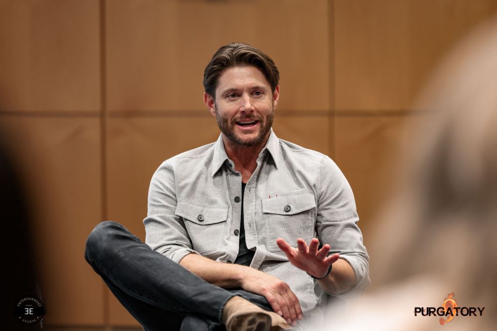 Jensen is sitting on a chair visible knees up. He is directly facing the camera while talking to someone to the right of the camera. He has a slight smile on his face while talking. He has his right leg over his left one with leaning with his ankle over left knee. His right arm is on his lap while he is mid movement with his left hand. Left upper arm is against his body while turned 90 degrees in front of him. Fingers splayed out and palm down.

He is wearing light brown shoes, black jeans, black shirt and a light beige button up jacket with chest pockets. His sleeves are rolled up. Background wall is rustic in color.