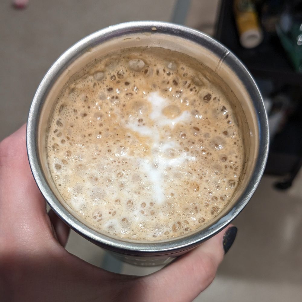 Top down view of a cappuccino/latte in a stainless steel travel cup. A hand is holding it and a thumb with black bail varnish is visible. The froth on the coffee almost looks like a tree with 5 branches.