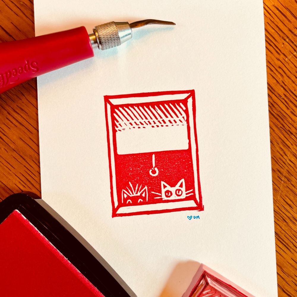 A small linocut stamp/print in red ink, of two cats peeking out a window, with a gouge tool, ink pad and the stamp block overlapping the paper.
