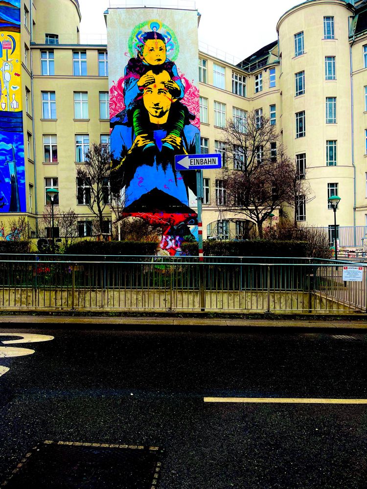 Nasser Asphalt. 
Quer eine Tiefgarageneinfahrt. 
Mittig ein Schild Einbahn. Pfeil Links. 
Dahinter ein Graffito an der Hausmauer. 
Mann nimmt Mädchen auf die Schultern. 