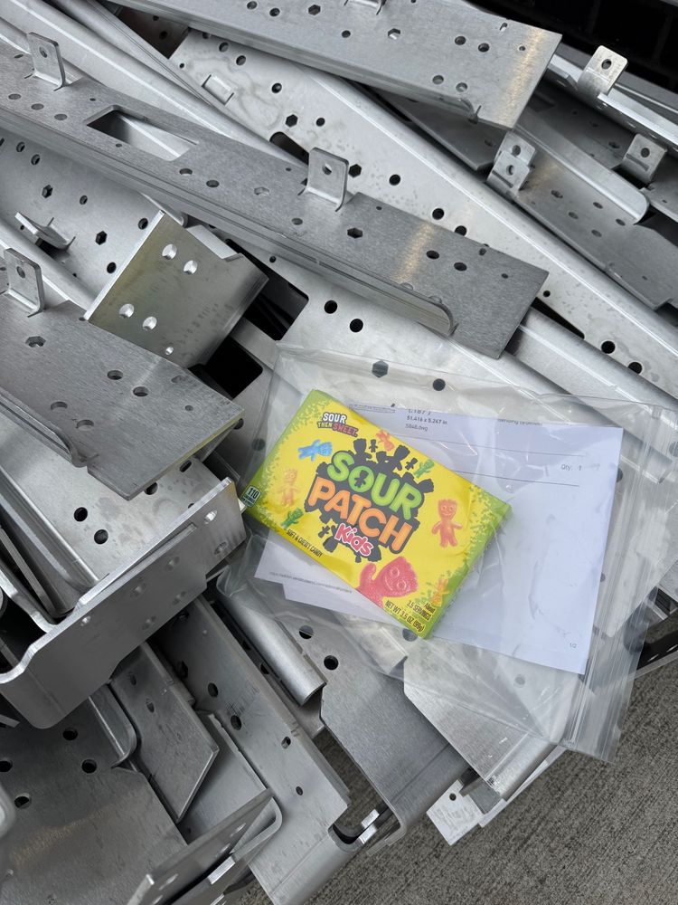 Close-up of a pile of metal parts, with a plastic bag that contains a receipt for the order, along with a box of Sour Patch Kids candies.