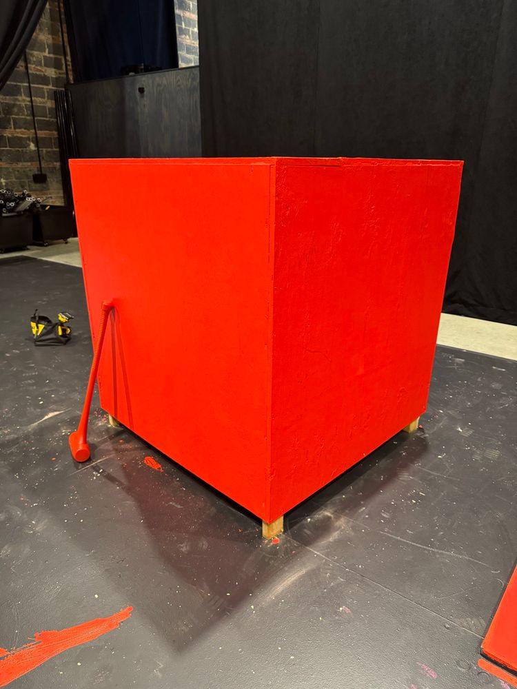 a very large red wooden box sitting on a black stage floor. a large red hammer leans against the box.