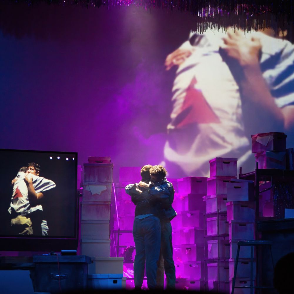 a production photo of a play named "the dance floor, the hospital room, and the kitchen table", showing two actors embracing amidst stacks of filing boxes