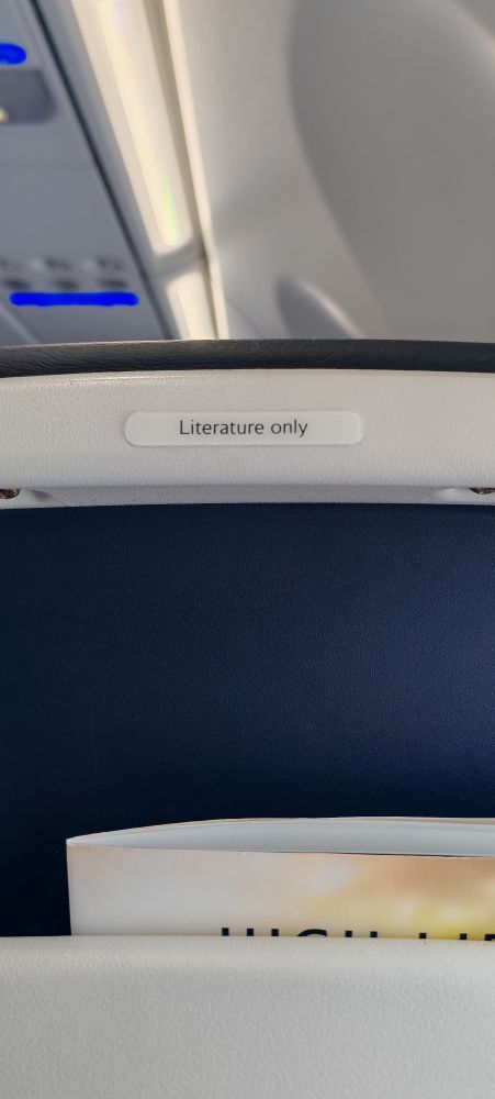 Photo of the back of a seat on an airplane, with a sign that says "Literature only" above a small alcove.