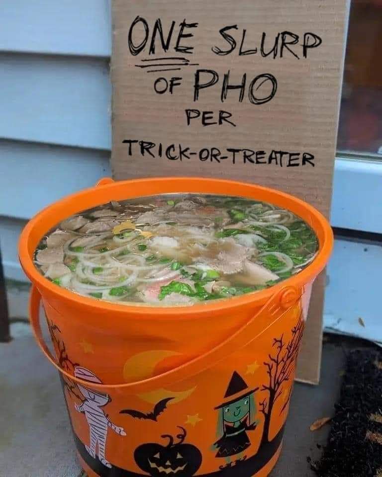 An orange Halloween candy bucket is full of soup. There is a hand written cardboard sign that reads "One slurp of pho per trick-or-treater"