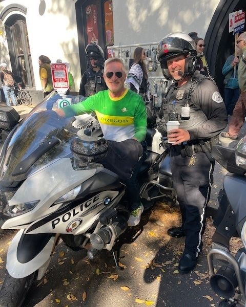 Dan Ryan sitting on a police motorcycle smiling, next to a cop with a coffee in hand.