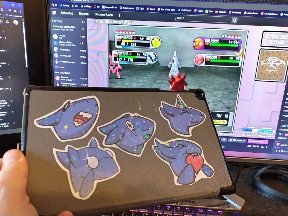 Image depicting holographic shiny stickers of the Sharky emote from Shauny7188's Twitch stream, made by Lupo Lycanth, applied to the back of a tablet, with Shaun's stream in that background playing Pokemon XD Gale of Darkness. 