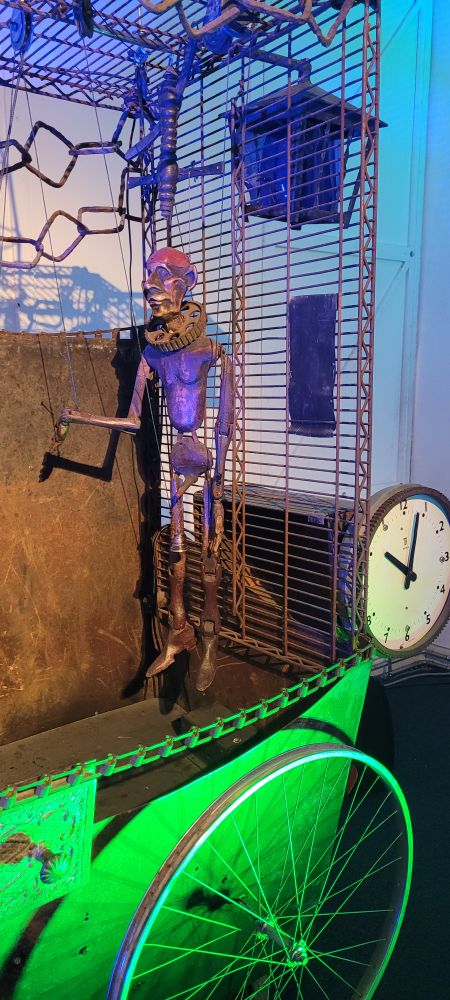 And image of a piece of kinetic art with a clock, wheel, cage and a man who has a bell at the end of his (ahem).