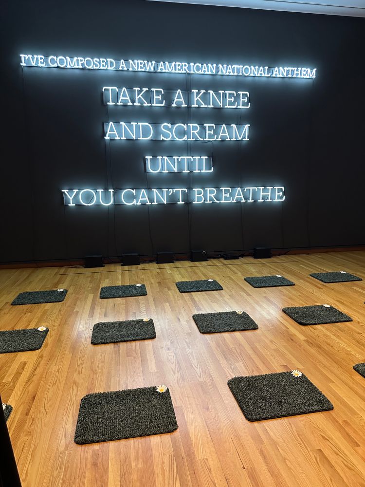 Astroturf mats on the floor in front of neon letters installed on a wall that say “I’ve composed a new national anthem: TAKE A KNEE AND SCREAM UNTIL YOU CAN’T BREATHE”