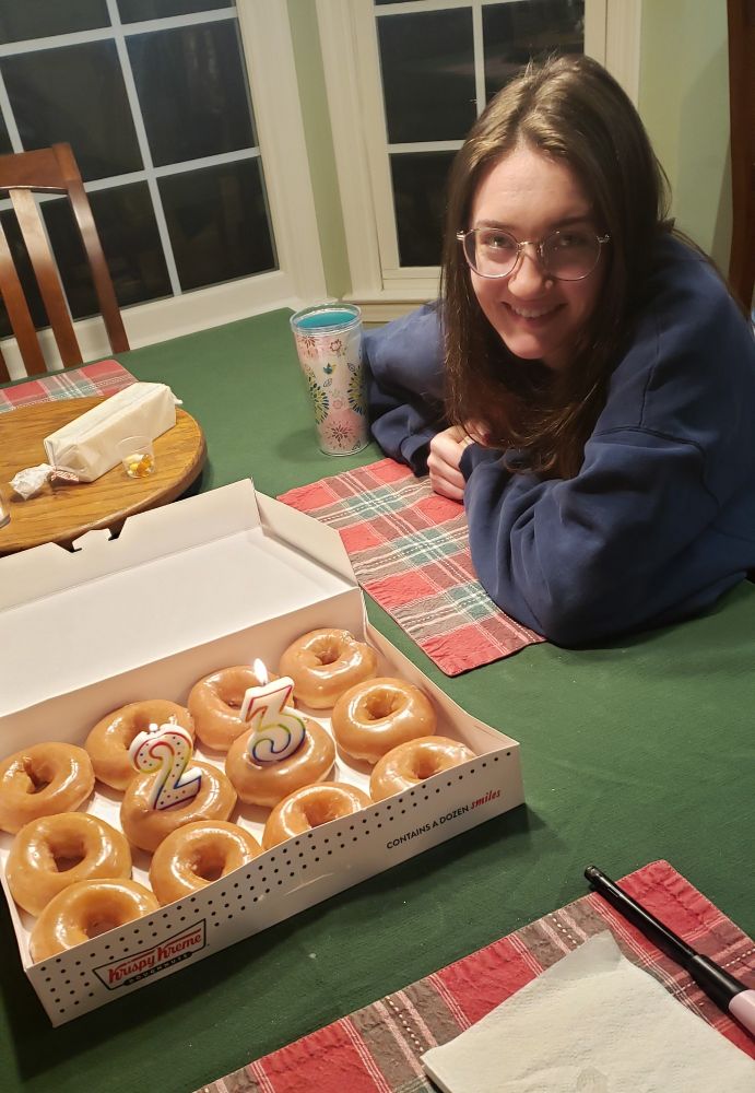 Young woman sitting at kitchen table next to a dozen Krispy Kreme doughnuts and a number 2 and number 3 candles lit. 