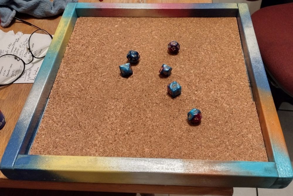 A large square wooden tray, spray painted with touches of bright colors, with a cork bottom and a set of dragon dice in the middle. 