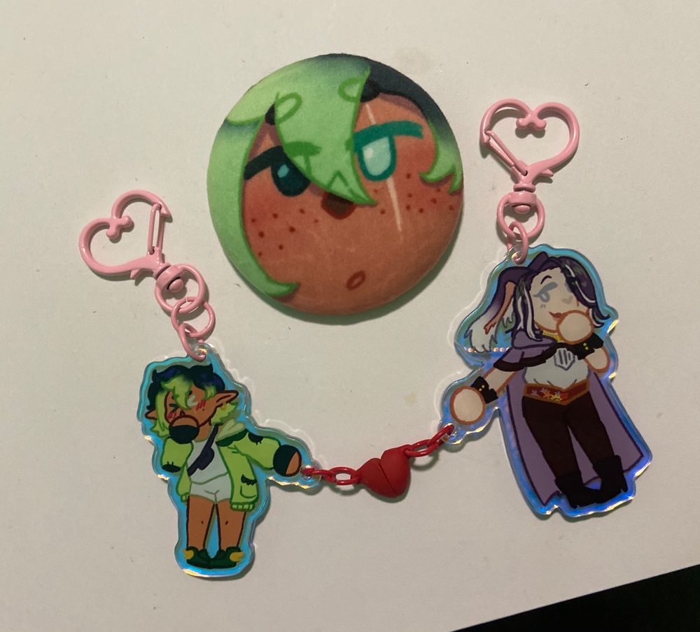 Three pieces of custom merch. One is a plush button, showing a close up of my wol’s face. Below it is two acrylic charms, connected by a magnetic heart. One is of my wol, a green themed lalafell. The other is my partner’s character, a purple themed viera. 