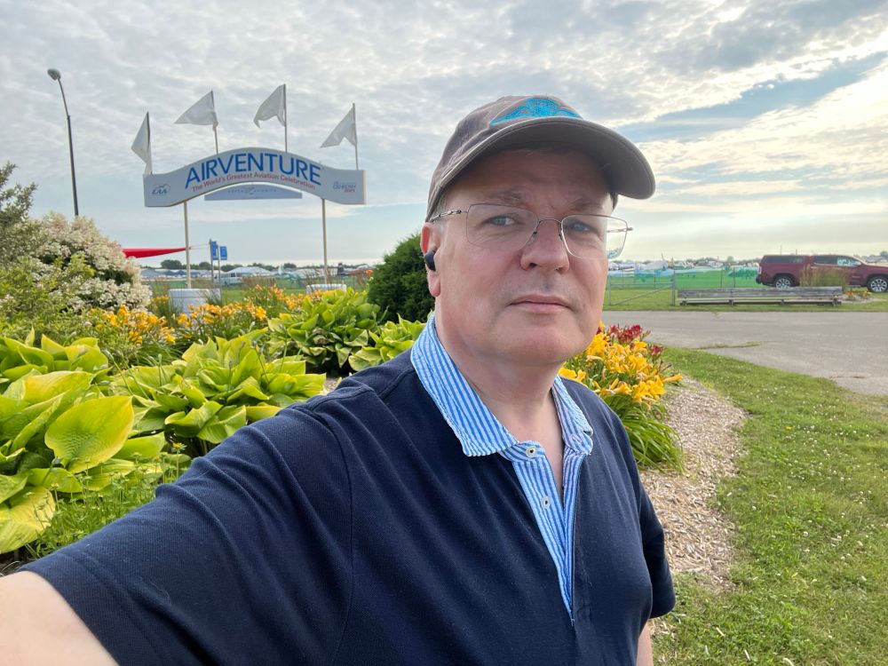 Me in AOPA ball cap standing in from of flowers and Airventure sign.