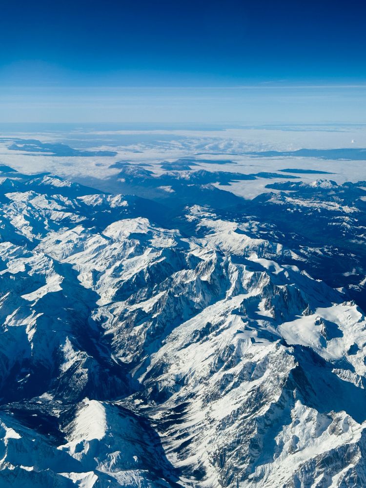 A view from an aircraft looking down over the snow-covered Alps, including Mont Blanc. The flatter land stretching in the distance towards the horizon. 