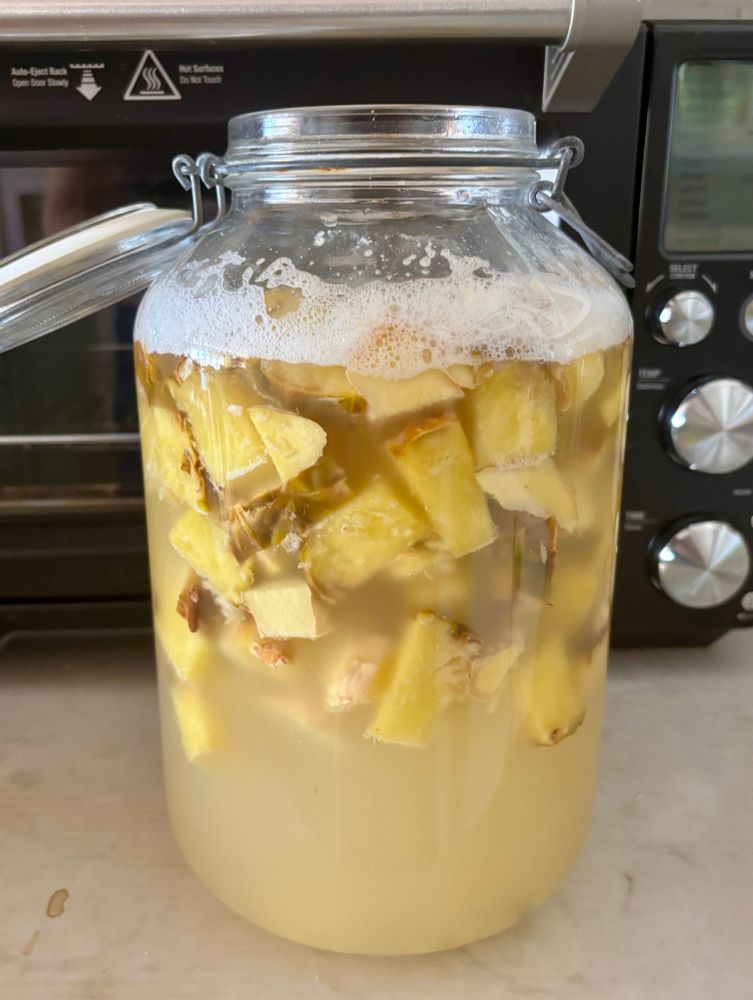 A large glass jar on a countertop filled with cubed pineapple, a light yellow liquid, and yeast bubbles (i.e. brewing tepache)
