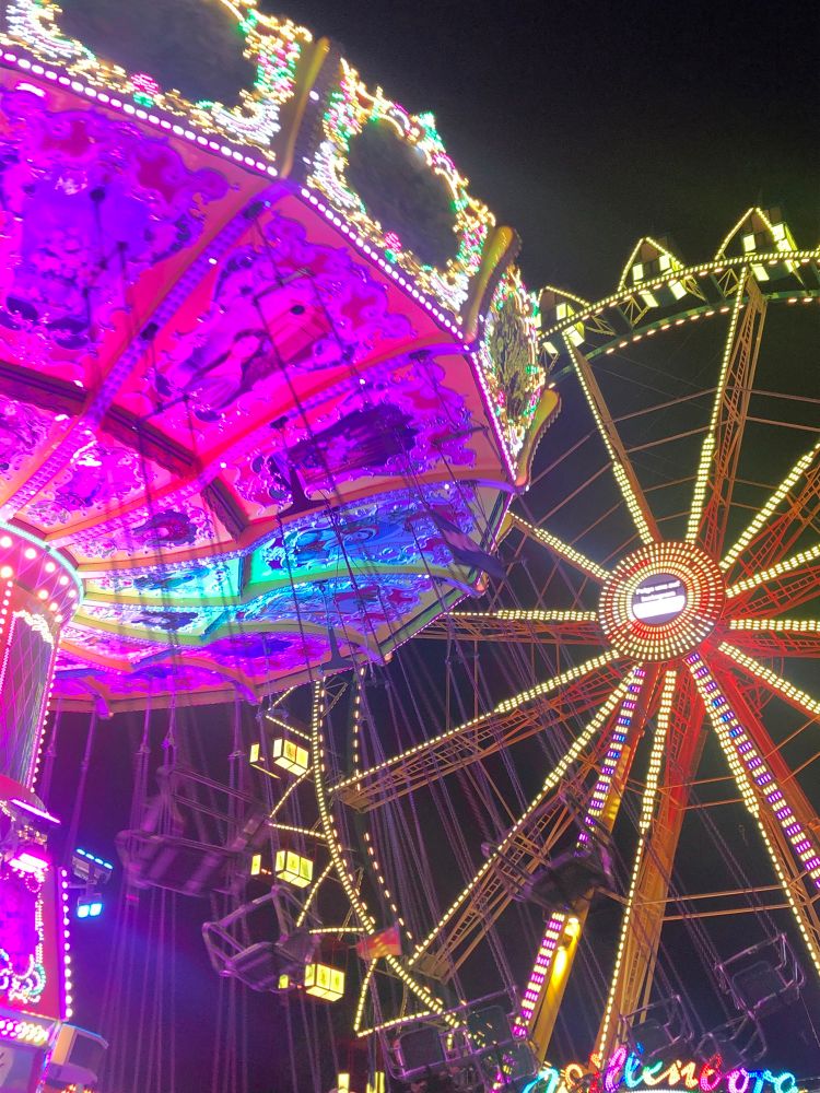 Karussell und Riesenrad auf einem Jahrmarkt. Abends, Dunkelheit, bunte Beleuchtung der Fahrgeschäfte. 