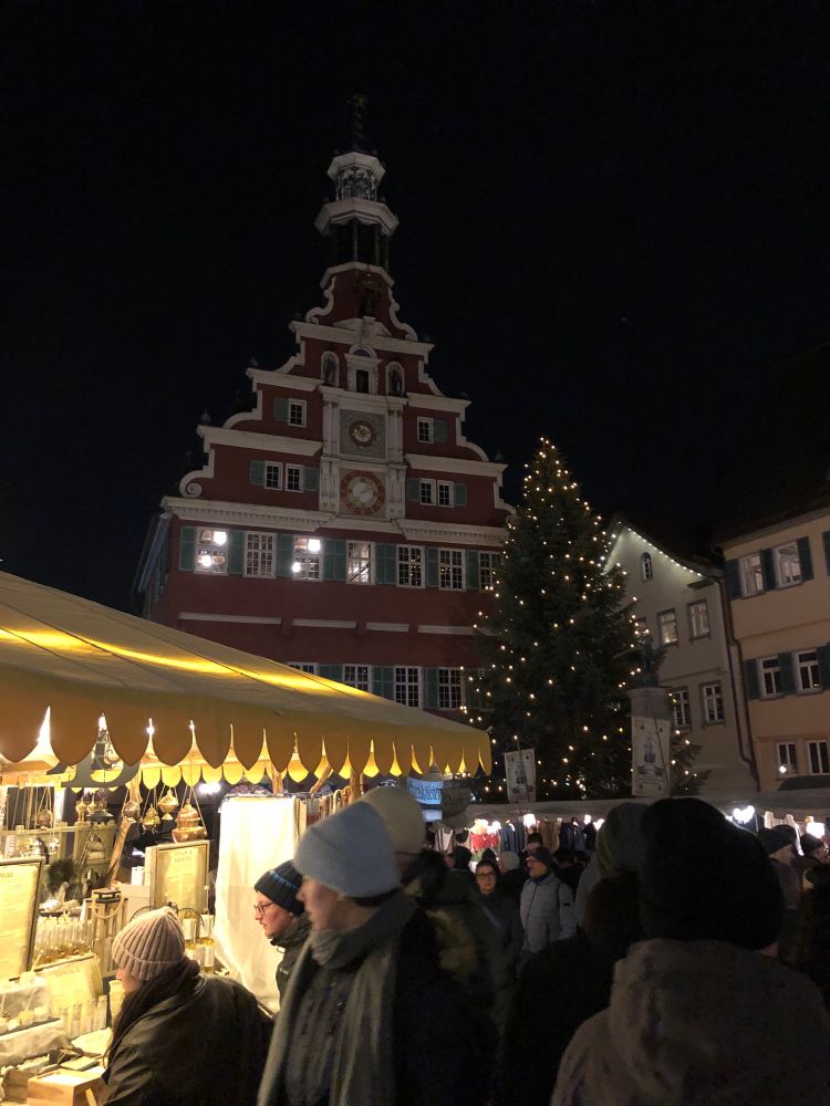 Das Bild zeigt einen Weihnachtsmarkt bei Nacht, dominiert von einem historischen Gebäude und einem geschmückten Weihnachtsbaum, inmitten einer Menschenmenge.