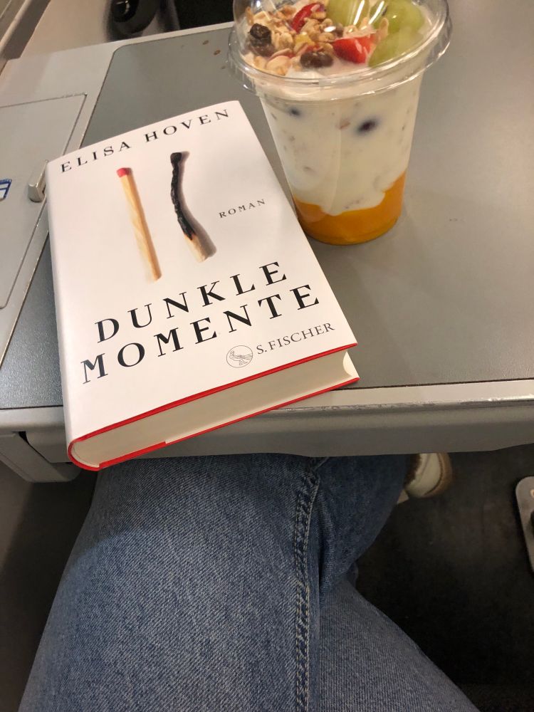 Auf einem Tisch im Zug liegt das Buch „Dunkle Momente“ von Elisa Hoven. Daneben steht ein To-go-Joghurt mit Obst. 