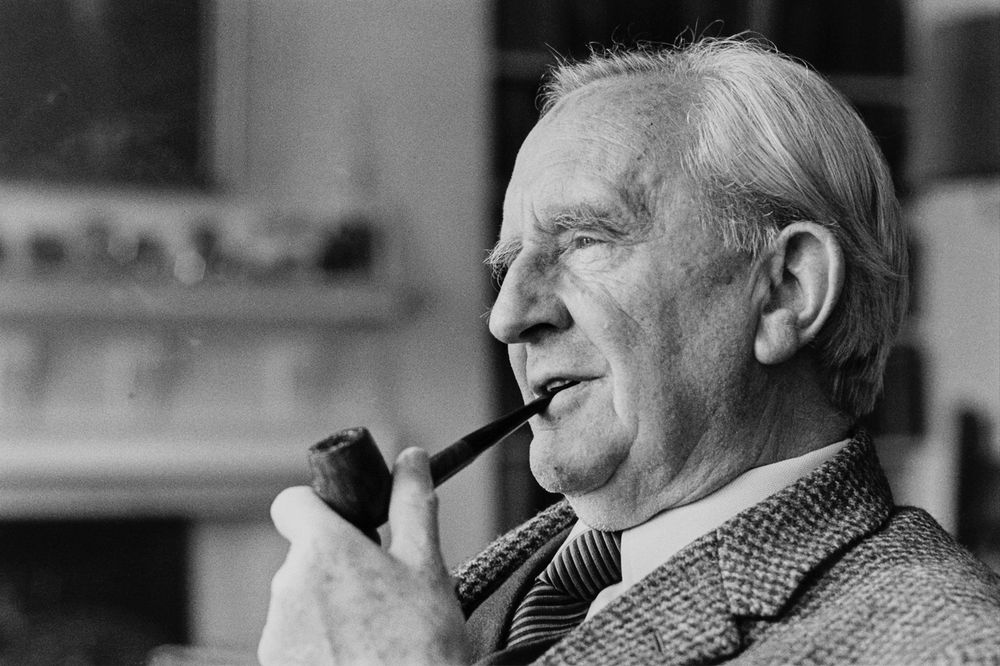 A black and white photo of an old Tolkien, with white hair. He's wearing a suit, smoking his pipe, with a light smile on his face.