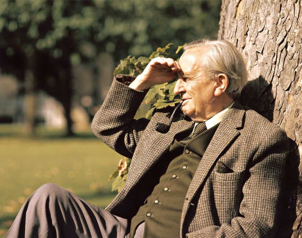 A colorized photo of an old Tolkien, with white hair. He's sitting down on grass, leaning on a tree, blocking the sun with his hand. He's wearing a suit and smoking his pipe.