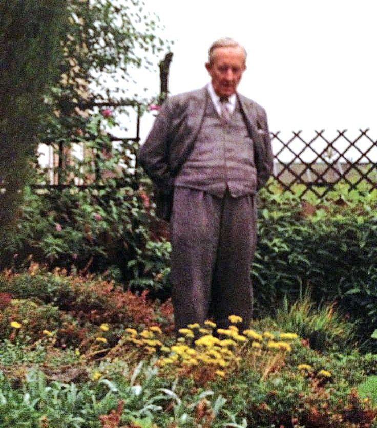 A colourized photo of an old Tolkien, with white hair. He's standing up in his garden, wearing a suit. He's looking down to some yellow flowers planted in front of him.
