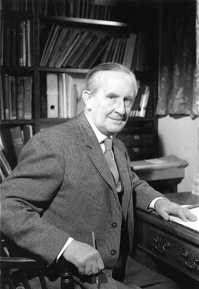 A black and white photo of an old Tolkien, with white hair. He's sitting in his office, wearing a suit. He's holding a pencil and looking directly into the camera with a soft smile.