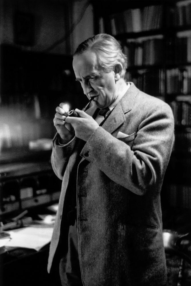 A black and white photo of an old Tolkien, with white hair. He's standing on his office, wearing a suite, lighting his pipe.