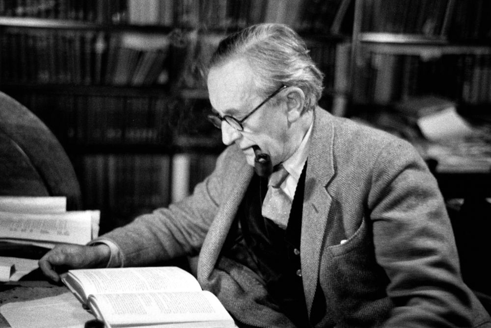 A black and white photo of an old Tolkien, with white hair. He's sitting in his office, wearing a suit and glasses. He's reading a book that's sitting on his table and smoking his pipe.