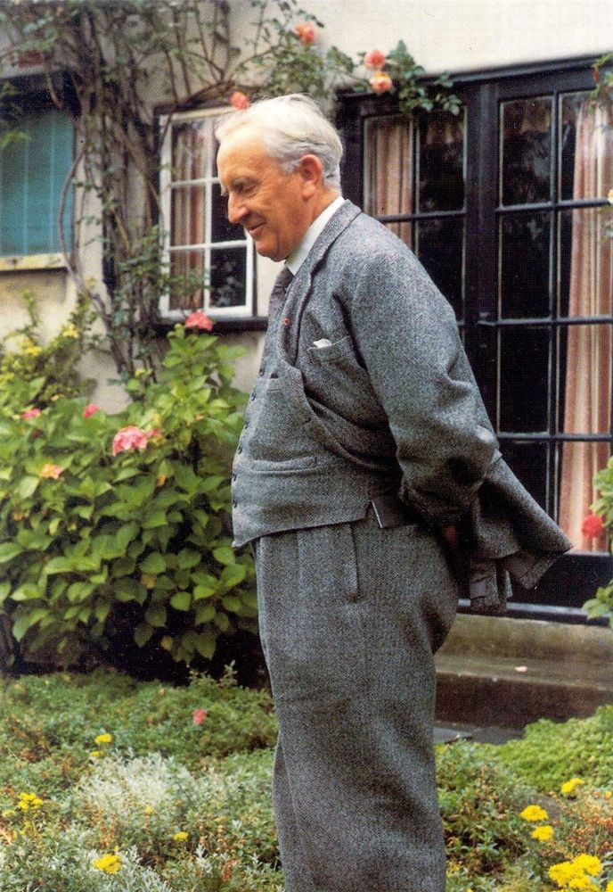 A colorized photo of an old Tolkien. He has white skin and a short white hair. He's wearing a grey suit, standing in a garden, with a lot of plants and flowers around him. His eyes are closed, he has a modest smile on his face and both his hand are on his back, under his suit.