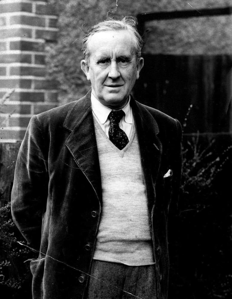 A black and white photo of an older J.R.R. Tolkien, with white hair. He’s standing outside, in a garden, dressed in a full suit, with his hands behind his back. He's looking straight to the camera with a soft smile.