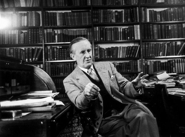 A black and white photo of J.R.R. Tolkien. He is older, with white hair. He is sitting in an office, surrounded by shelves and desks overflowing with books. He is wearing a full suit and gesturing with his hands, as if speaking to someone not visible in the photo. He is white, has short hair neatly combed close to his head, and is clean-shaven.