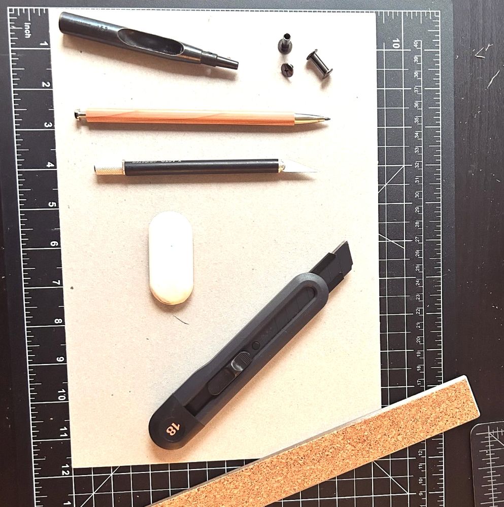 Gazing down on materials for making book art: a black self healing cutting mat, a metal ruler cork-side up, a black utility knife, white oval eraser, black xacto knife, wood barrel mechanical pencil, black steel hole punch, and black chicago screw posts, on 8x11 book board.
