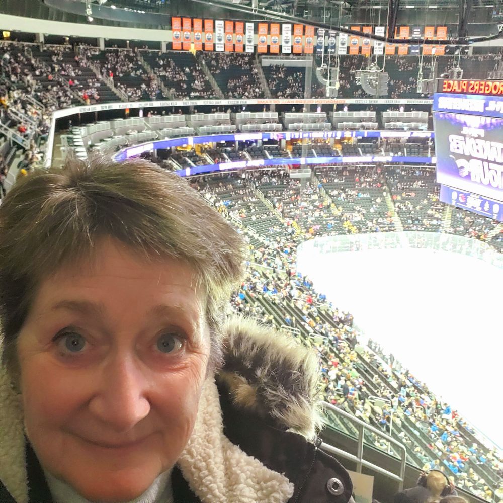 A woman in the foreground, high up in the stands, with the lower seats and ice rink behind her in the distance. 