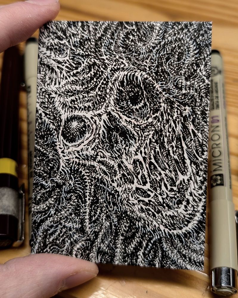 Photo of a sketch card illustration with pens in the background. The card's illustration is a detailed black and white ink depiction of a horrific face surrounded by tendrils.