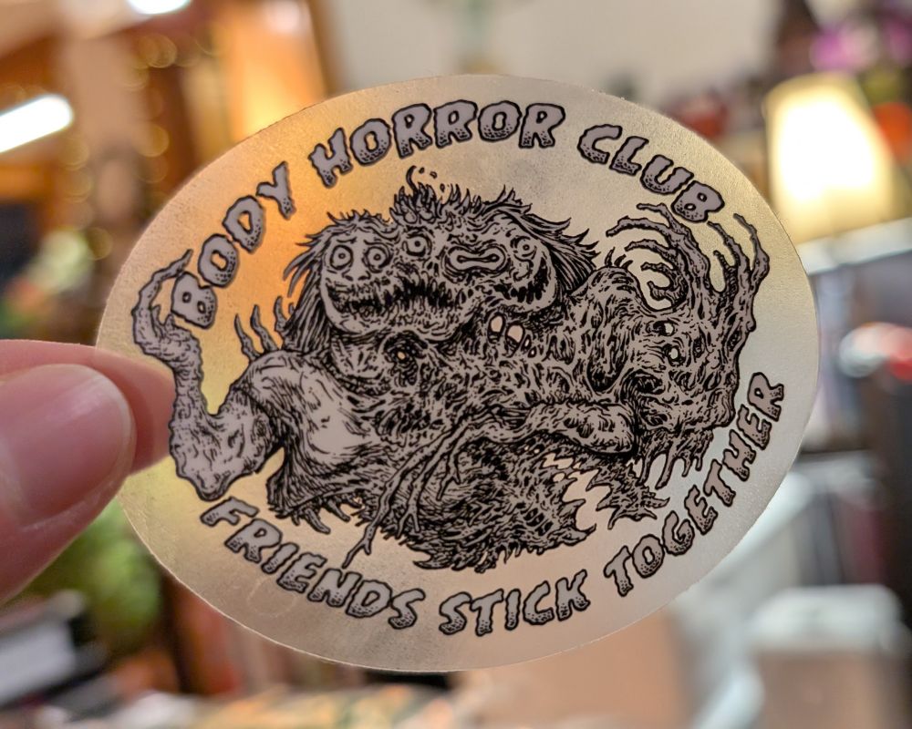 Picture of sticker being held up over a blurred background, the sticker features a black and white illustration on a transparent background that allows light to show through the parts that aren't colored by the white or black or the art. The art is a gruesome mass of the upper bodies of three people stuck together with twisted flesh and sharp teeth all around. The people are smiling, like they're having the time of their lives. The text at the top, above the body mass, reads Body Horror Club and the text below reads Friends Stick Together, all text is in a stylized font in all caps. 