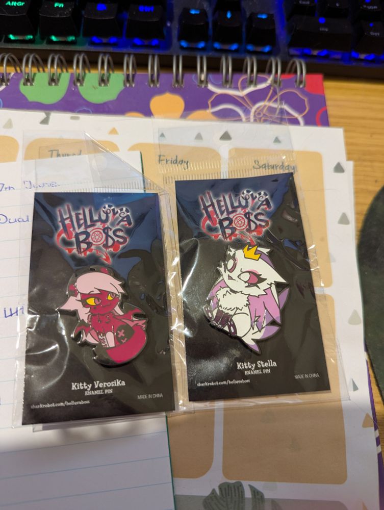 Two Helluva Boss kitty pins, one featuring Verosika and one featuring Stella, still in their plastic and backing on a desk pad. 