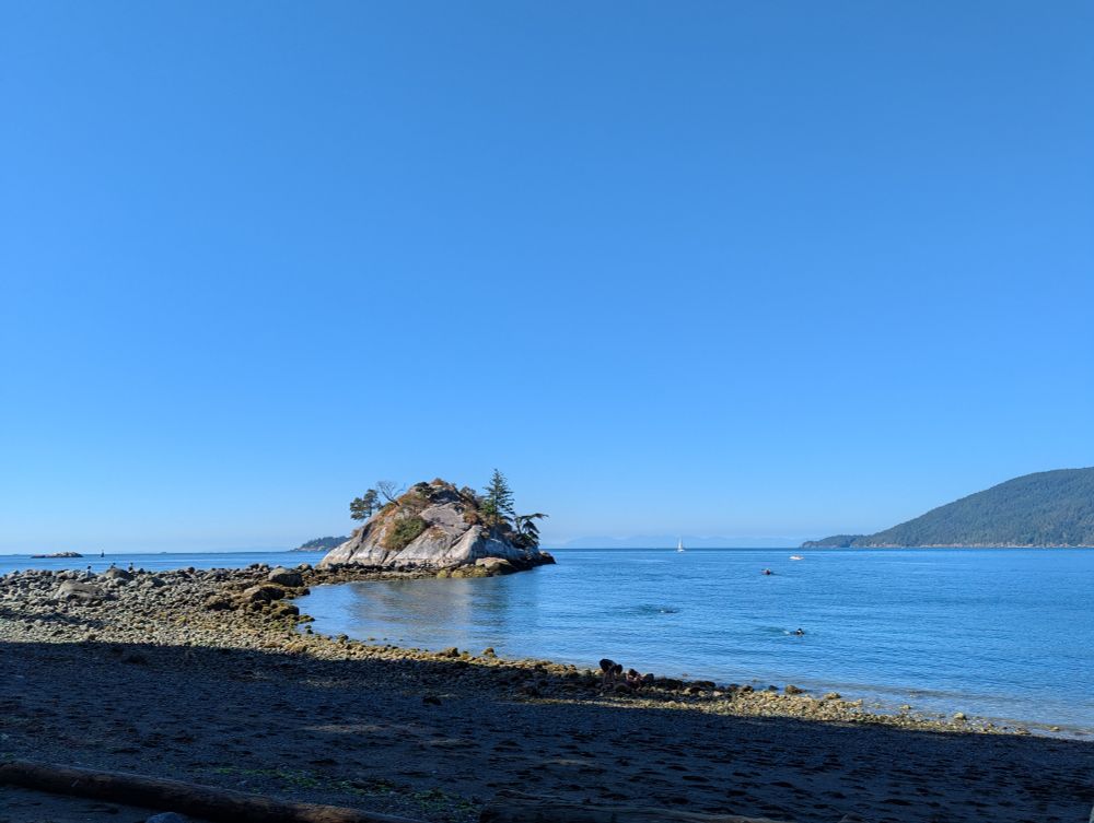 Photo of a rocky beach with calm blue ocean under a pure blue sky. The beach extends out to a peninsula with large rock on it, covered with scraggly trees and moss, worn smooth by the elements. Tiny people are visible climbing the sloped rock face.