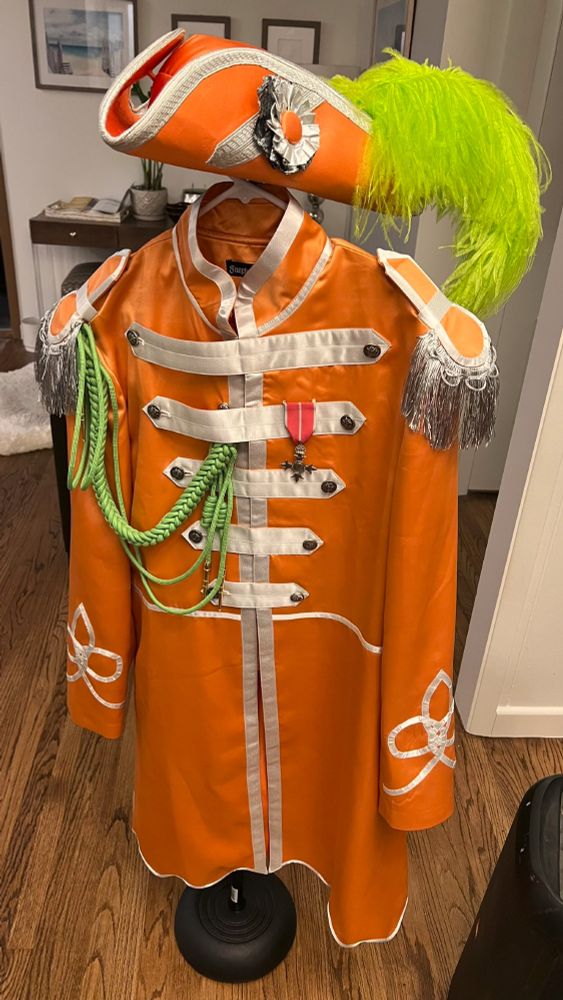 An orange and white marching band jacket with a green shoulder cord and a matching orange and white tricorn pirate hat with a lime green feather hanging on a microphone stand.
