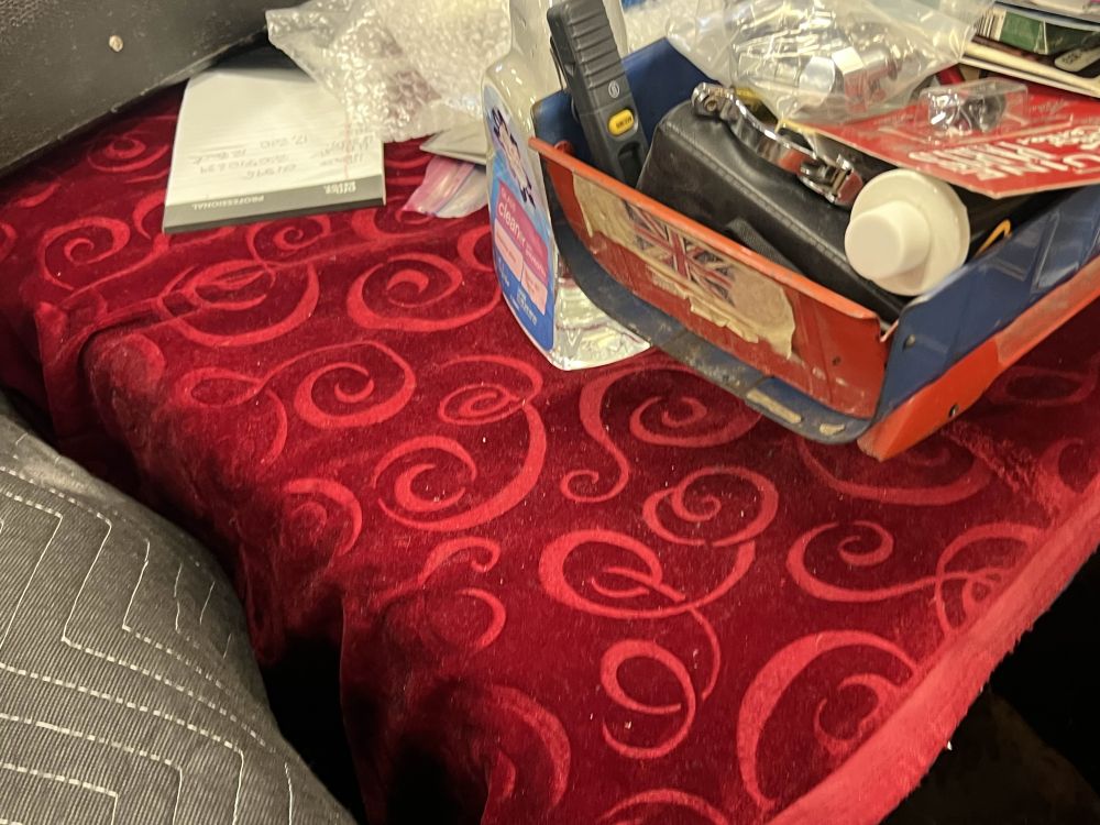 A swatch of red velvet with an abstract swirl pattern. There is a box of random supplies, a spray bottle, and bubble wrap on top of it.