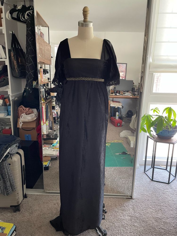 A photo of a black velvet dress with short sleeves and gold trim under the bust line and lace-edged sleeves. The dress is on a dress form in a messy room, with a window in the back and a money plant in the corner. 
