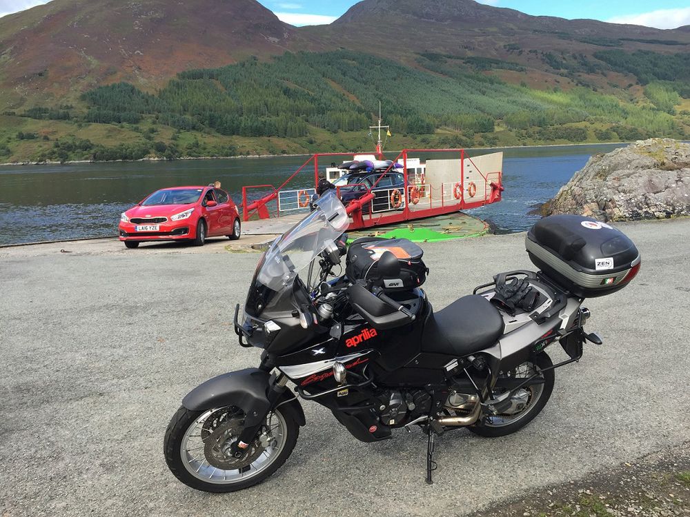 Large black motorcycle in the foreground while red and black cars drives off off a small ferry behind it 