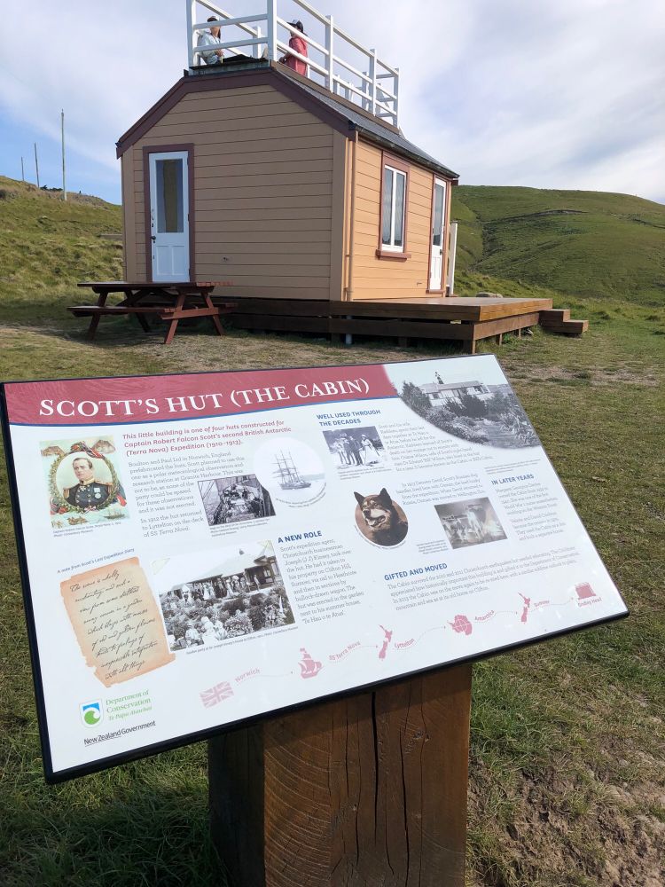Plaque in front of small wooden cabin on hillside. Plague explains Scott’s cabin’s travels from England to Antarctica & to Aotearoa New Zealand.