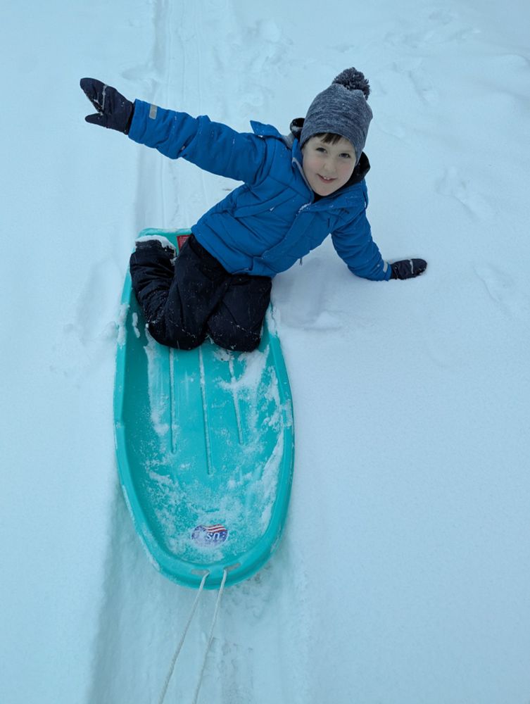 Photo of a happy boy in a blue jacket and snow pants, on a teal sled. He has his right arm up and his left hand is in the snow.