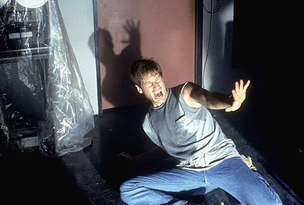 Man in jeans and grey sweat covered t shirt on the ground holding up his hand with a terrified face. 