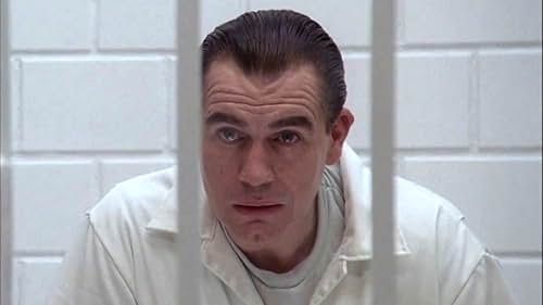 A shot of a seated man through prison bars. He is dressed in white, which matches the bright white of the bars and brickwork in the cell. His black hair is slicked back, and he is leaning forward with an expression of casual but absolute contempt on his face, as if he were toying with something far beneath his intellectual abilities.