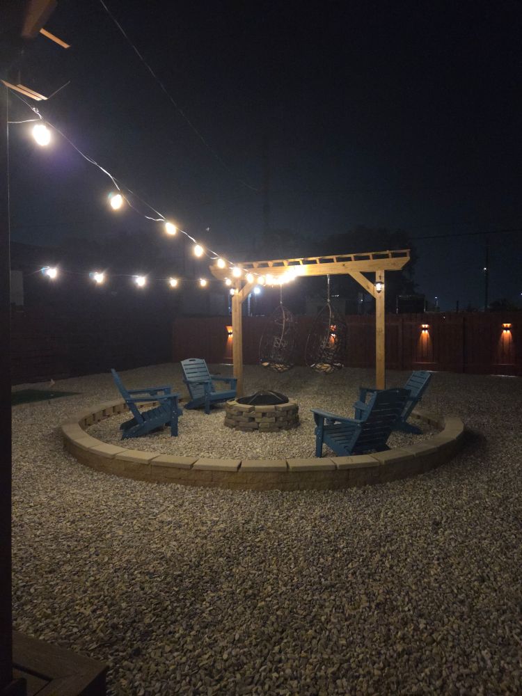 A fenced in yard at night with party lights strung to a tellus. 2 hanging chairs are hung from the trellis, joined by 4 lawn chairs circling a fire pit. The ground is covered in gravel.