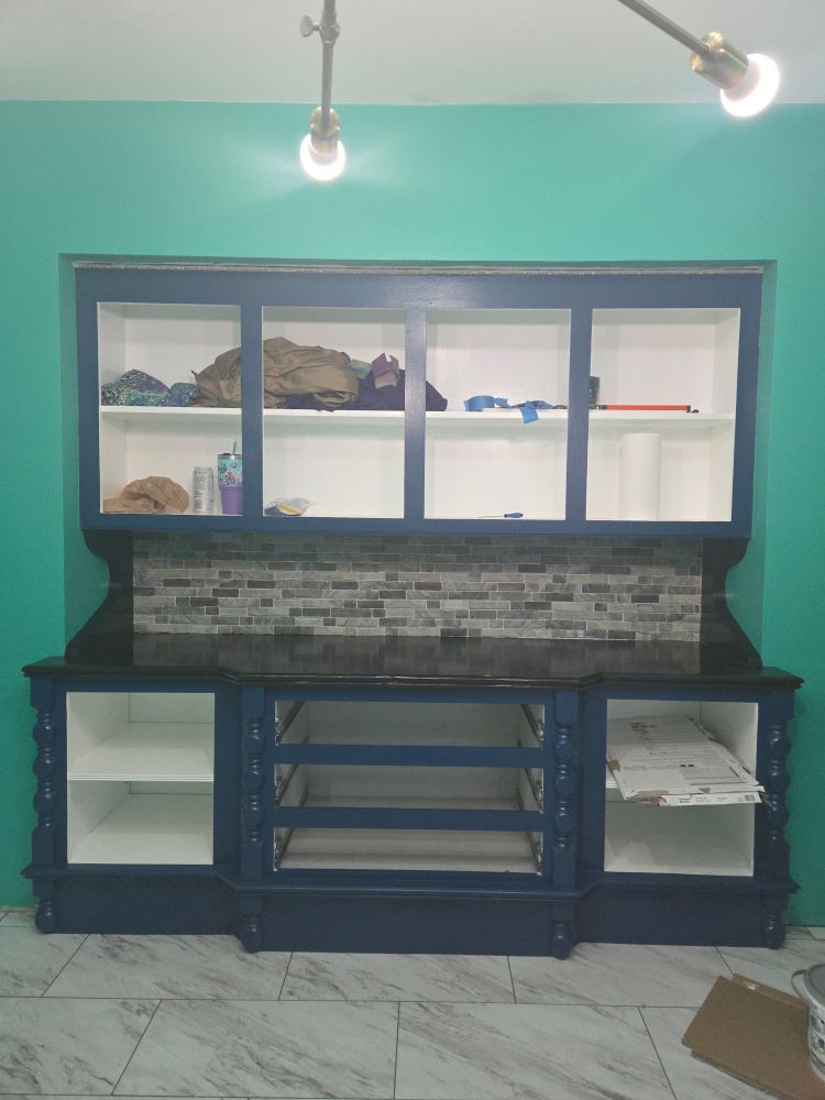 A green wall has a vintage built in cabinet with the door removed. The inside is painted white, the outside is painted navy blue, and the countertop is stained black with a glossy topcoat. A backsplash with a mix of gray patterned ceramic and glass tiles.