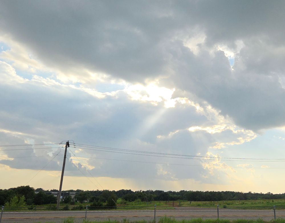 An Oklahoma skyskape of clouds and sunbeams shining through over the highway, trees, and plains.